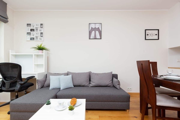 A cozy living room featuring a gray sectional sofa, a small white coffee table, and wall-mounted artwork. The space is illuminated by natural light from a nearby window.