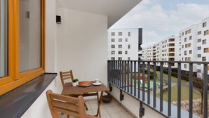 Un balcón con mesa y sillas de madera, una planta pequeña en maceta y una ventana con marco amarillo.