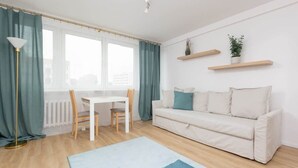 Una sala de estar moderna con un sofá blanco, una mesa de comedor y sillas. Hay dos ventanas con cortinas, una lámpara de pie y dos estanterías con plantas.