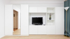 Una sala de estar moderna con un televisor de pantalla plana sobre un mueble blanco, una planta pequeña en un estante y una lámpara de pie.