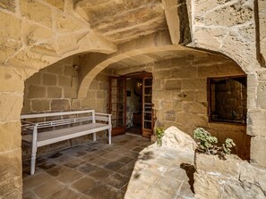 A stone-walled courtyard with a bench, an open door leading to another room, and a small plant on a rock.
