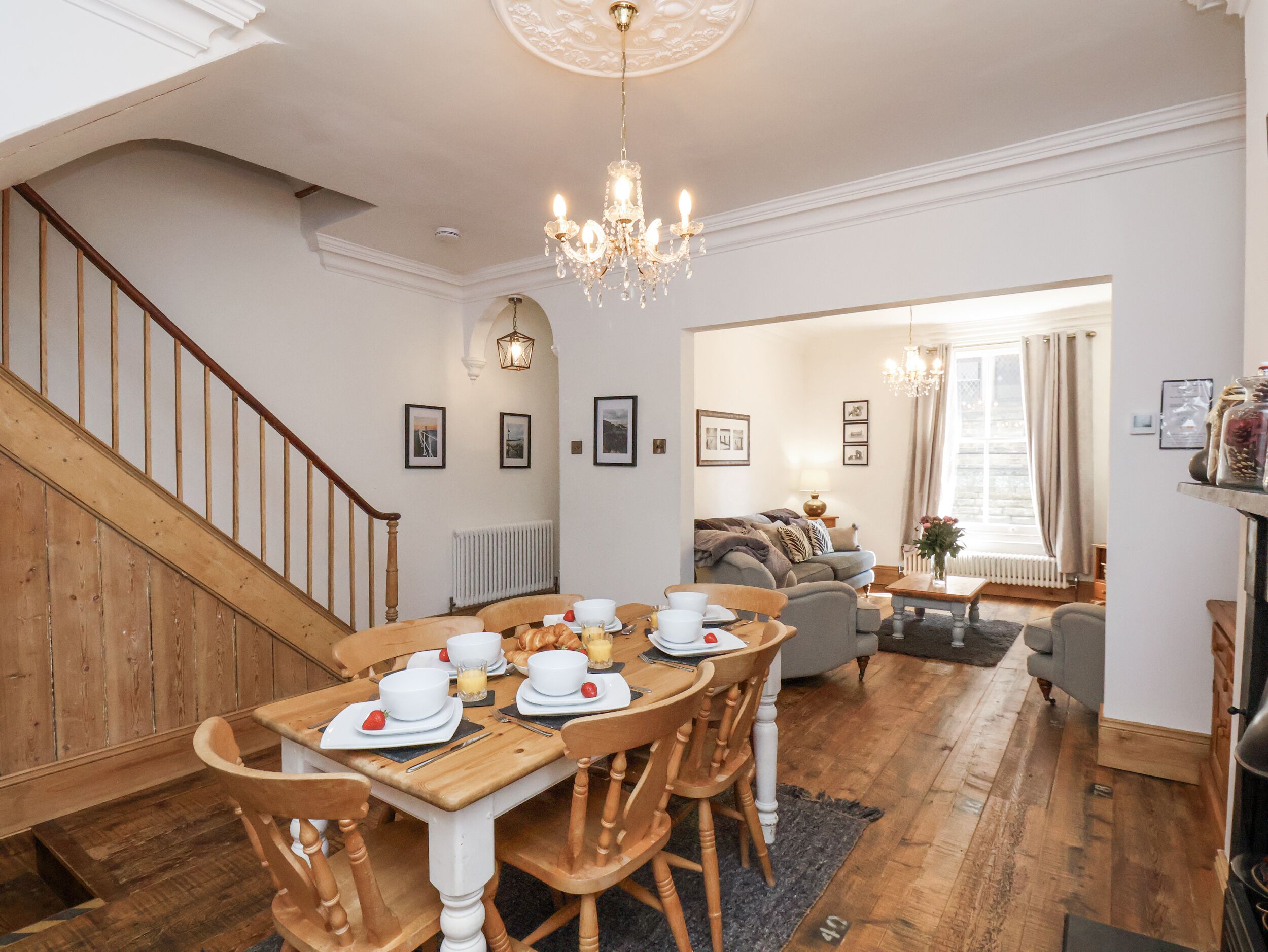 Comedor con una mesa de madera preparada para cuatro, una escalera y una sala de estar con sofá y ventanal.