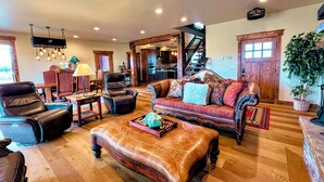 A spacious living room with a leather sofa, a matching ottoman, and a dining area with a wooden table and chairs. A kitchen is visible in the background. - Luxury Family Home with Full Teton Views + Hot Tub (Driggs)