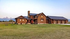 A large, rustic two-story house with a prominent chimney, multiple garage doors, and a spacious front lawn. - Luxury Family Home with Full Teton Views + Hot Tub (Driggs)