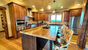 A spacious kitchen with wooden cabinets, stainless steel appliances, and a large island with bar stools. - Luxury Family Home with Full Teton Views + Hot Tub (Driggs)