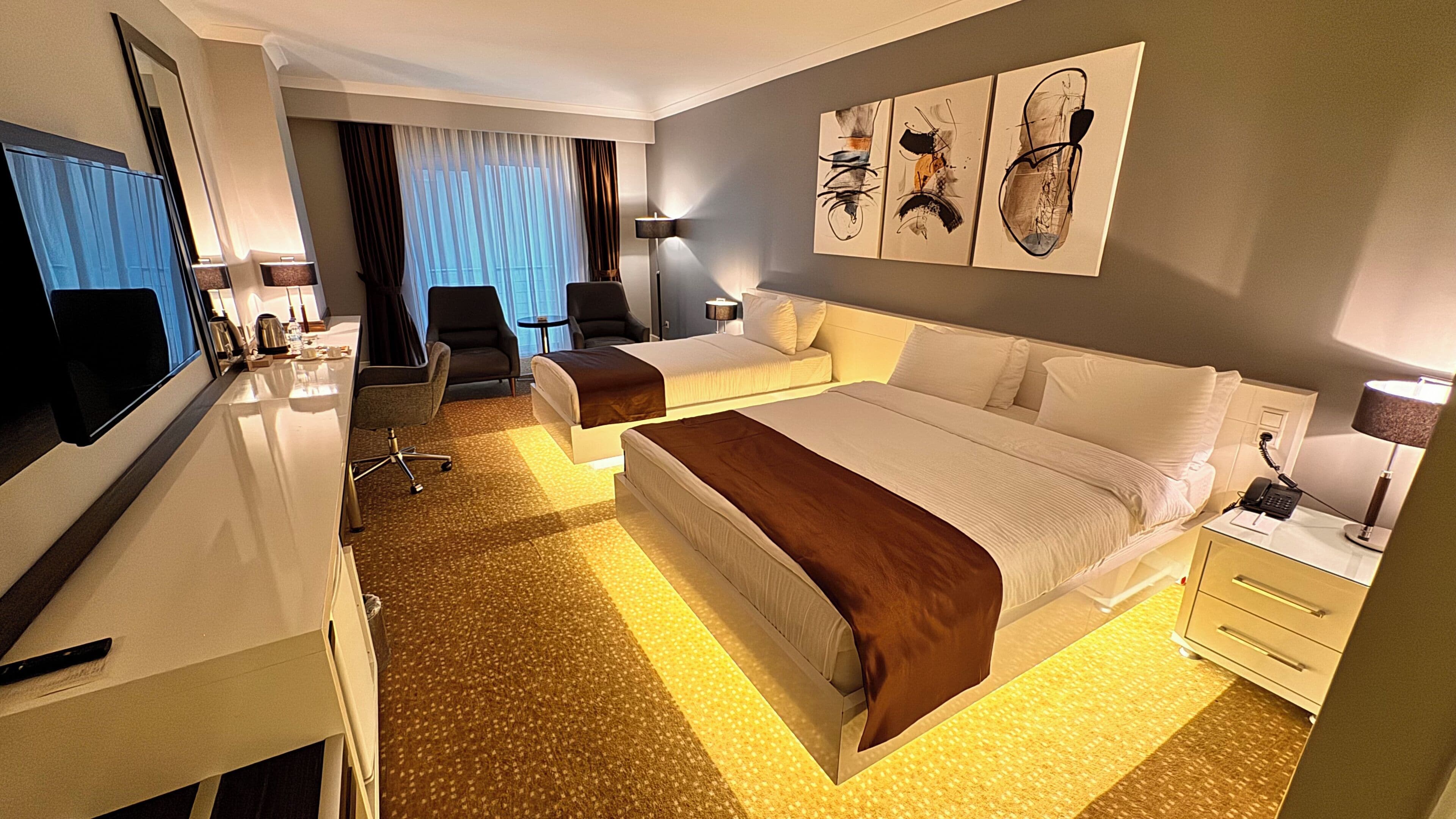 A hotel room with a bed, two chairs, a desk, a mirror, a lamp, and artwork on the wall.