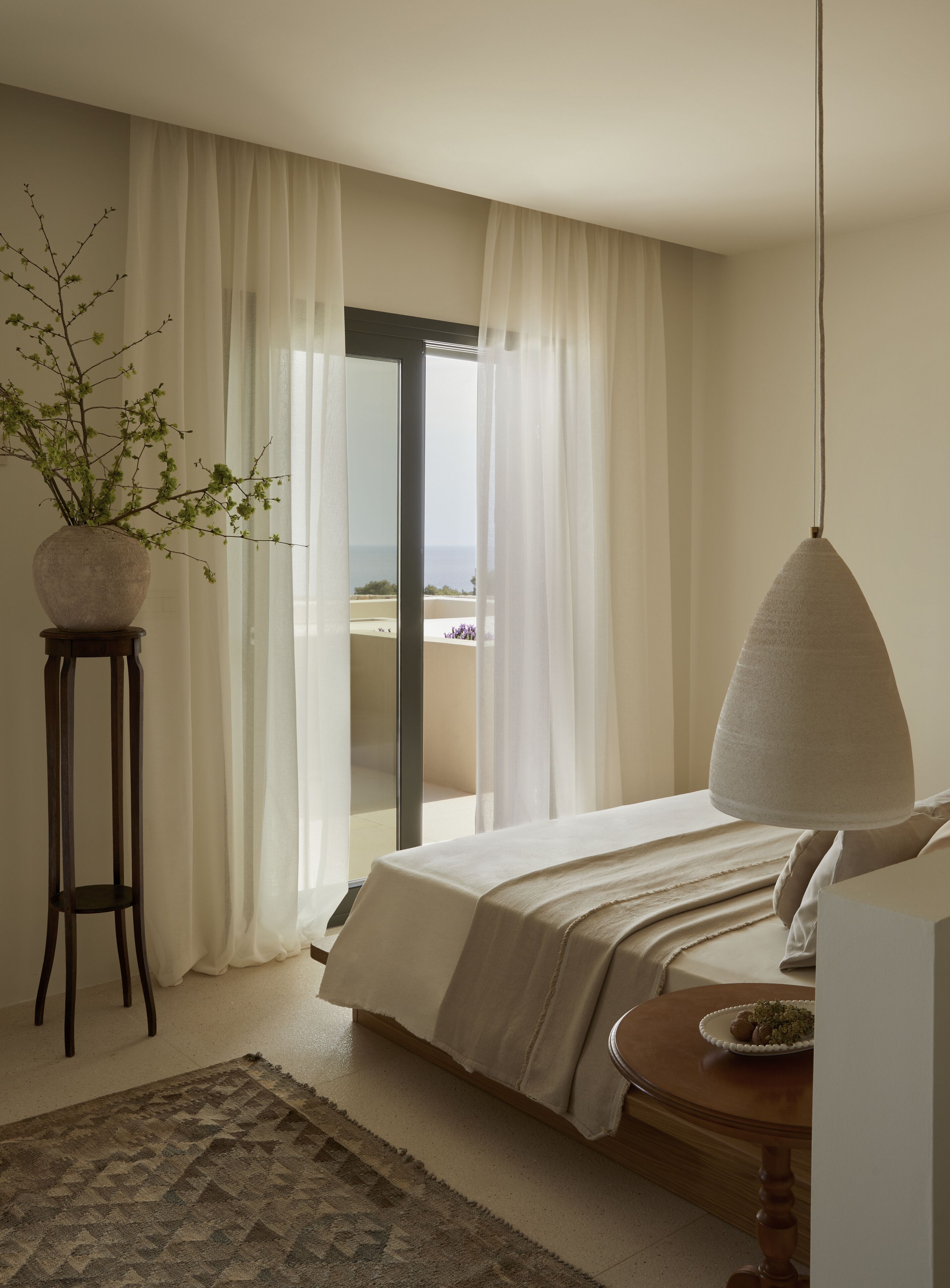 A bedroom with a bed, a bedside table with a lamp, a potted plant, and a view of the sea through a large window.