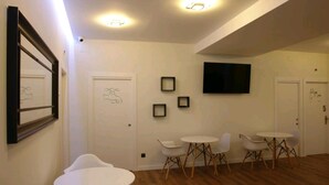 A modern room with white walls, a flat-screen TV mounted on the wall, and a wooden floor. There are two white round tables with chairs, and a door with a number '5' on it. - Hostel Trovadores (Burgos)