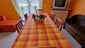 Et spiseområde med et ternet borddække, træstole, en blomsterdekoration i midten og et fjernsyn på et stativ.