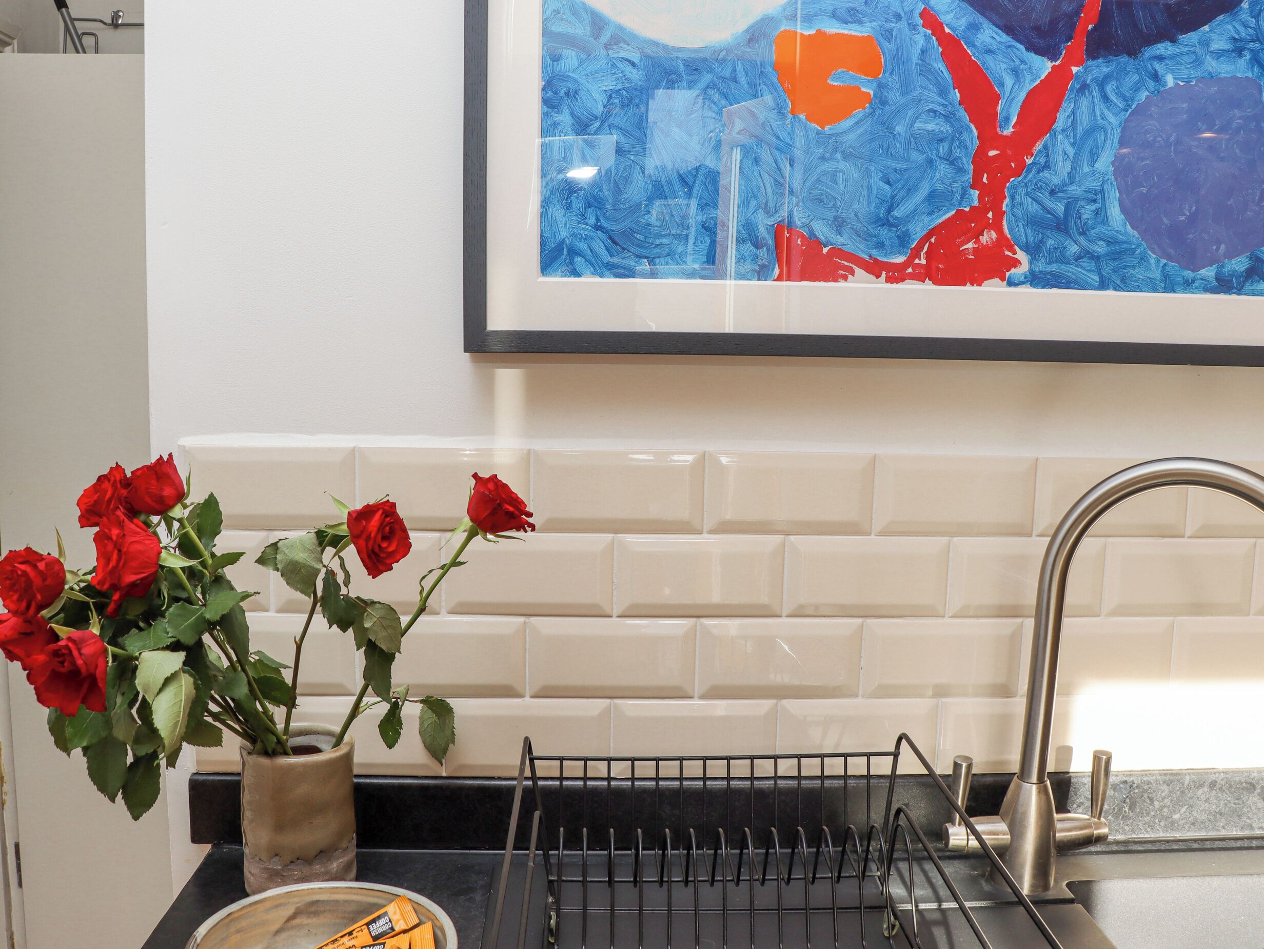A kitchen with a white tiled backsplash, a stainless steel sink, a black countertop, and a vase with red roses.