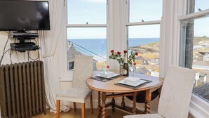 A room with a large window overlooking the sea, a table with chairs, a television, and a radiator. - Whistler (St Ives)