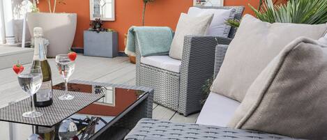 A wicker sofa with cushions, a small table with a bottle and glasses, and potted plants.