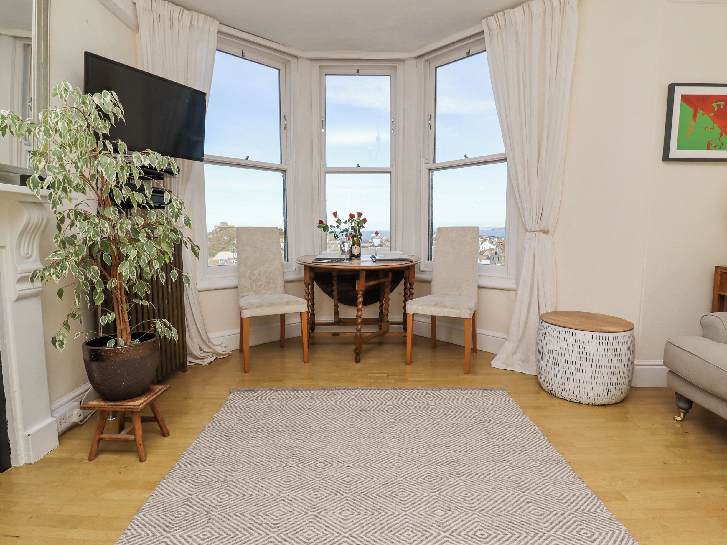 Um quarto com janela panorâmica, uma mesa de jantar com cadeiras, um vaso de planta, uma mesa lateral redonda e um sofá.