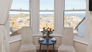 A bay window with two chairs and a table, overlooking a coastal town with buildings and a hill. - Whistler (St Ives)