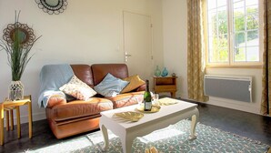 A cozy living room with a brown leather sofa, a white coffee table, and a patterned rug. There is a window with curtains, a wall-mounted mirror, and a small side table with decorative items. - Vouvray - Beautiful castles (Vouvray)