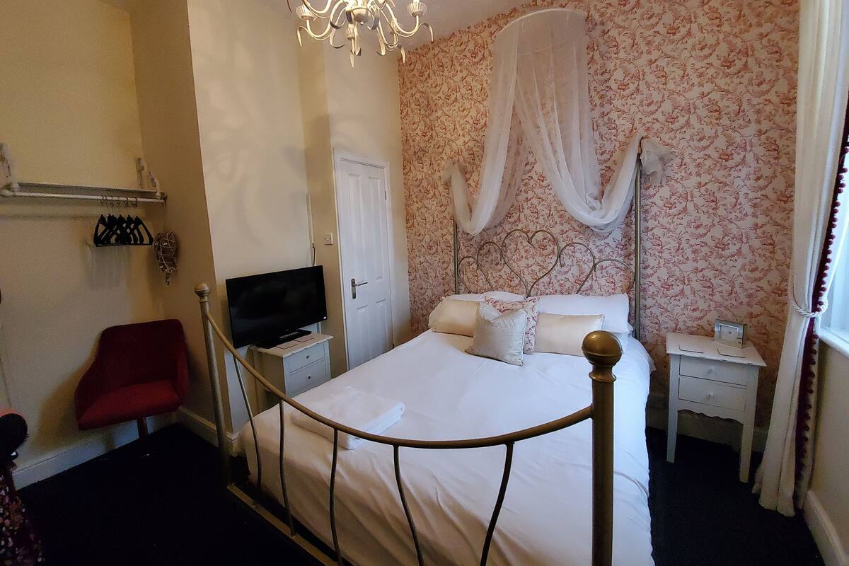 A bedroom with a bed, a television, a red chair, a nightstand, and a chandelier.