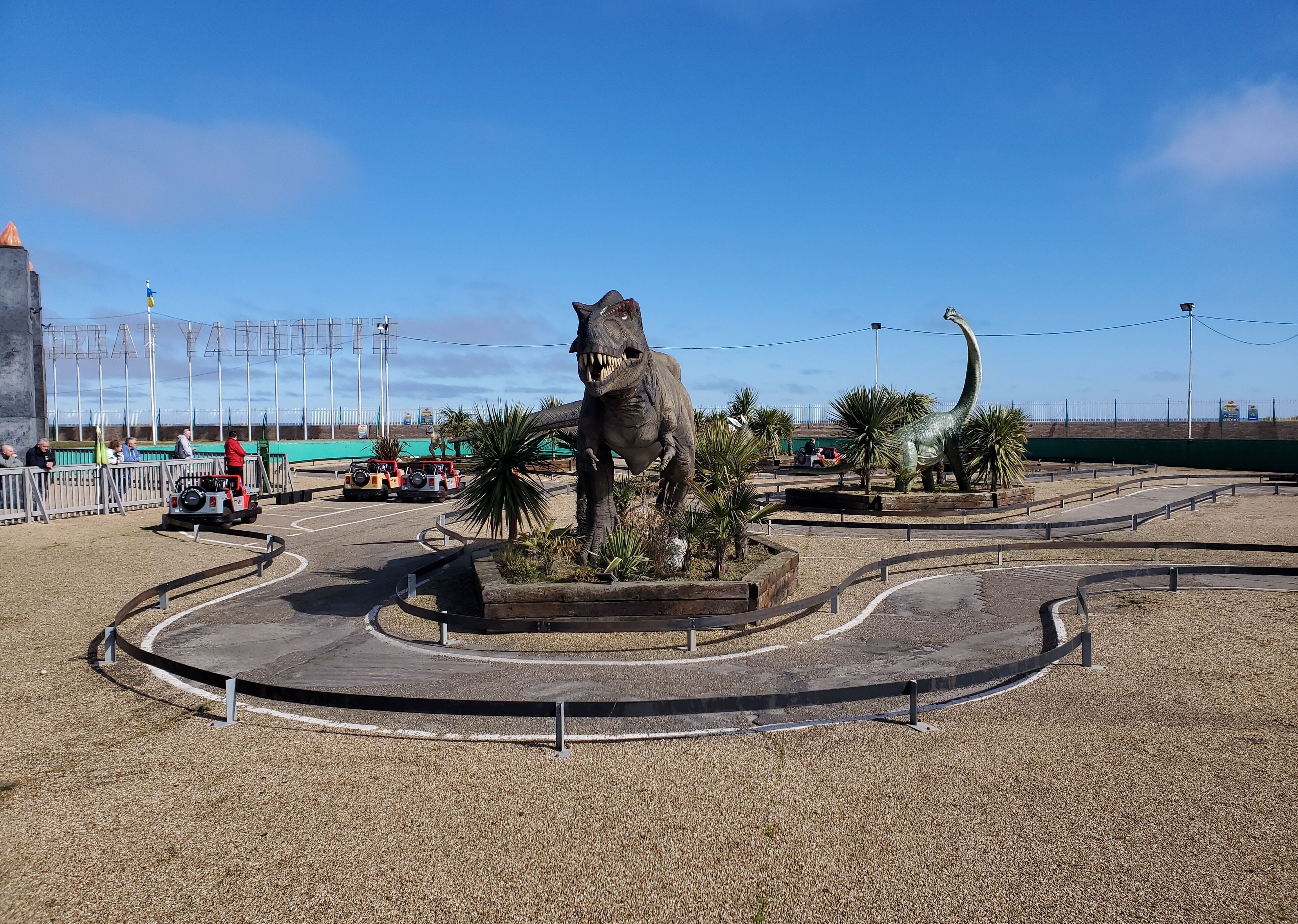 A dinosaur statue in a landscaped area with a paved path and a fence.