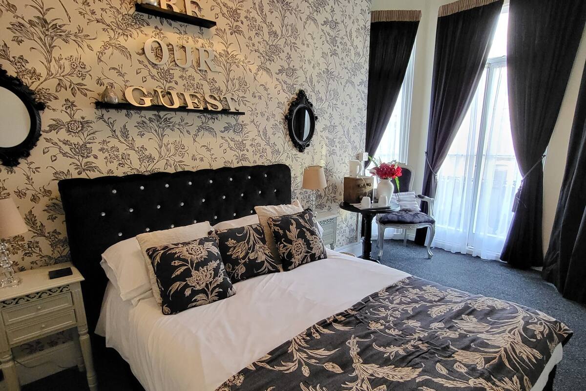 A neatly arranged bedroom with a large bed, a bedside table, a mirror, and a wall-mounted shelf with decorative items.