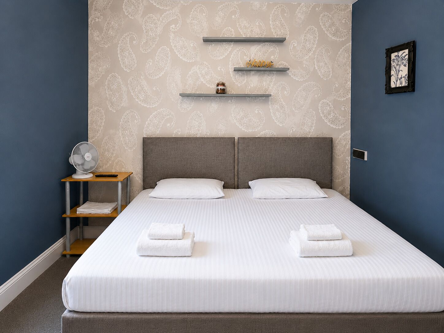 Cama doble con ropa blanca y dos almohadas, mesita de noche con un ventilador y una pared con estampado de paisley.