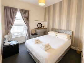 Et hotelværelse med seng, skrivebord med lampe, spejl, vindue med gardiner og en radiator.