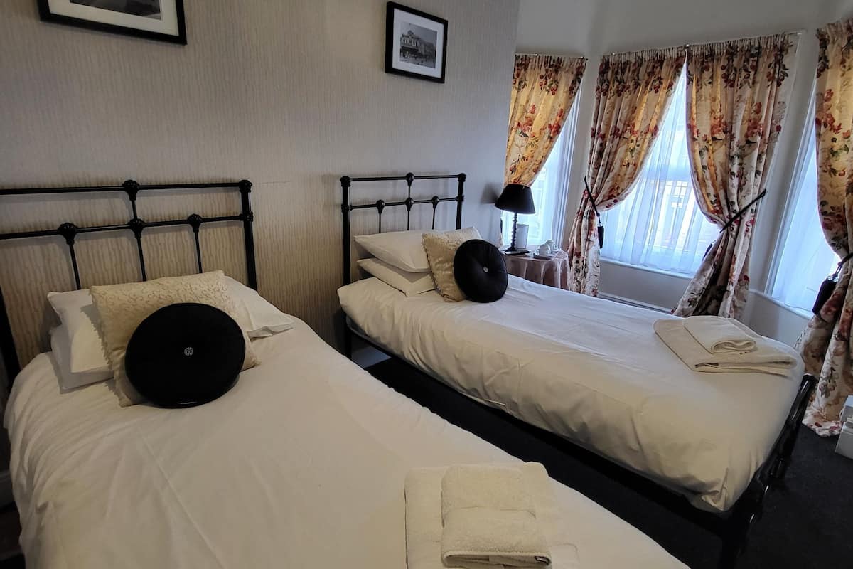 A hotel room with two beds, each with white linens and black headboards. There are two framed pictures on the wall, a window with floral curtains, and a bedside table with a lamp.
