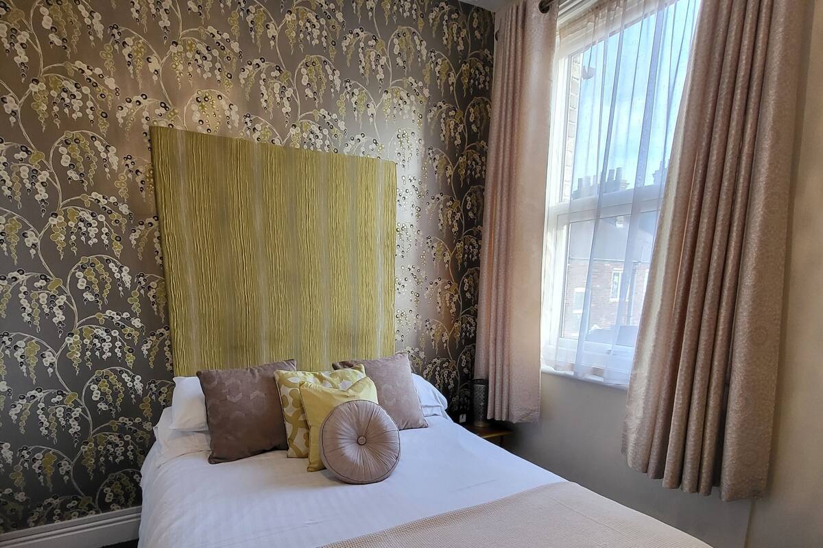 A neatly made bed with a textured cover, a headboard with a vertical pattern, and a window with sheer curtains.