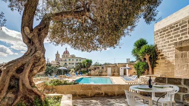 A pool area with a table and chairs, a large tree, and a building with a dome in the background. - Dar ta' Lulu - 2 bedroom house with valley views (L-Ghasri)