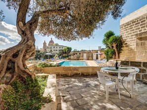 A pool area with a table and chairs, a large tree, and a building with a dome in the background. - Dar ta' Lulu - 2 bedroom house with valley views (L-Ghasri)