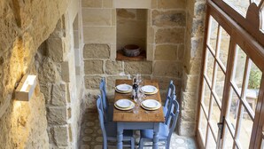 A stone-walled dining area with a wooden table set for four, blue chairs, and a view through a large arched window. - Dar ta' Lulu - 2 bedroom house with valley views (L-Ghasri)