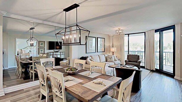 A spacious living area with a dining table, chairs, a sofa, and a balcony with large windows. - Sandpiper CRD1C - Beautifully Decorated - Private Stairs to pool and beach (Pawleys Island)