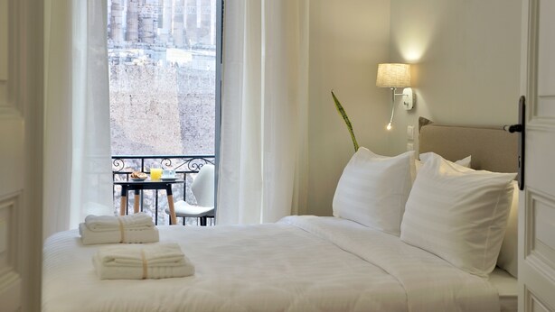 A hotel room with a bed, a nightstand with a lamp, a window with a view of buildings, and a balcony with a table and chairs. - 1900 Athenian House (Athens)