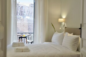 A hotel room with a bed, a nightstand with a lamp, a window with a view of buildings, and a balcony with a table and chairs. - 1900 Athenian House (Athens)