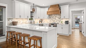 A modern kitchen with white cabinetry, a large island with stools, a herringbone-patterned backsplash, and a view of a pool through an open door. - 🏖Steps 2 Beach Access🏴‍☠️Pirate Themed🏝️Private Pool/Luxury/Location (Miramar Beach)