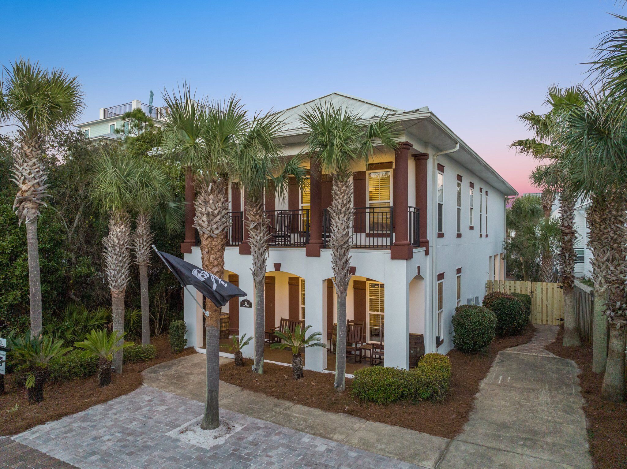 A two-story house with a balcony, surrounded by palm trees and a paved driveway.