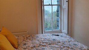 A bedroom with a floral bedspread, a radiator, and a window with a view of trees. - Impeccable 1-bed Apartment in Ilfracombe (Ilfracombe)