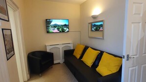 A compact living space with a black sofa, yellow cushions, a white door, and two mounted TVs displaying outdoor scenes. - Impeccable 1-bed Apartment in Ilfracombe (Ilfracombe)