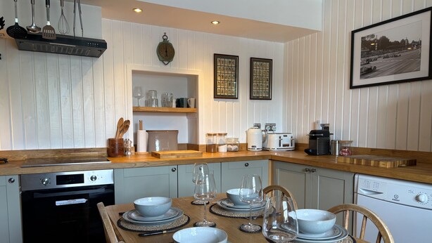 A compact kitchen with modern appliances, a dining table set for six, and a framed photo on the wall. - 1 Keepers Cottage, Skeabost Bridge, Isle Of Skye (Skeabost)