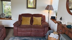 A living room with a brown leather sofa, two cushions, a lamp, a framed picture, a radiator, a window with blinds, a small table with decorative items, and a basket with towels. - 1 Keepers Cottage, Skeabost Bridge, Isle Of Skye (Skeabost)