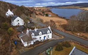 A white house with a blue door, surrounded by a rural landscape with a lake and hills. - 1 Keepers Cottage, Skeabost Bridge, Isle Of Skye (Skeabost)