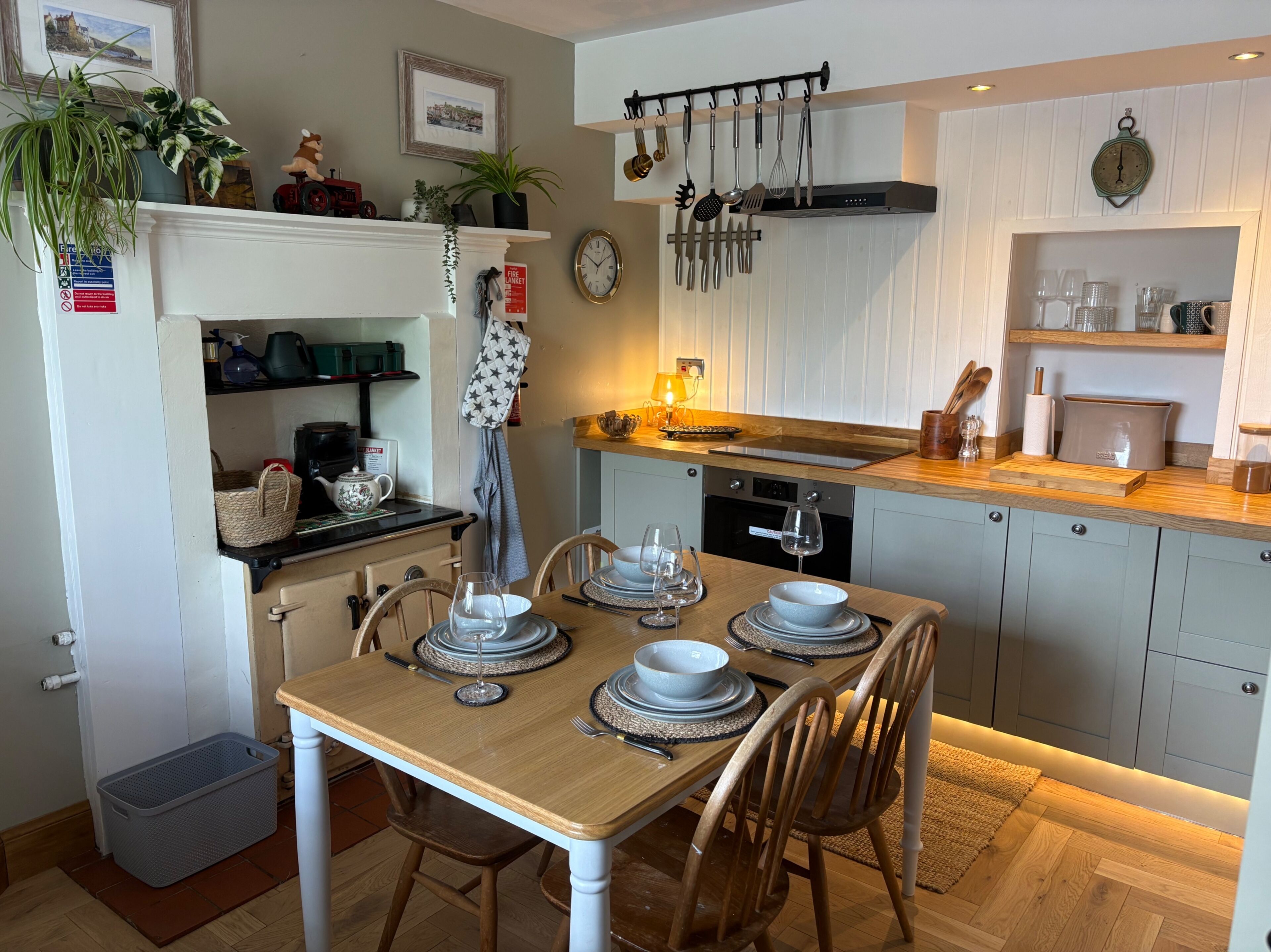 A dining area with a wooden table set for four, a kitchen with a stove, oven, and hanging utensils, and a small shelf with decorative items.
