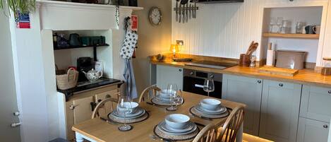 A dining area with a wooden table set for four, a kitchen with a stove, oven, and hanging utensils, and a small shelf with decorative items.
