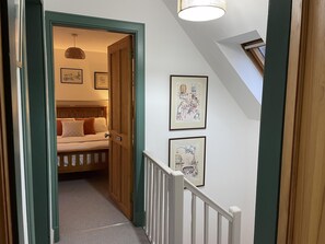 Corredor com porta de madeira que dá para um quarto, onde se vê uma cama e um lustre. Quadros emoldurados decoram a parede.