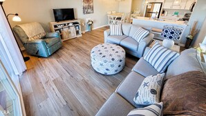 A modern living room with a sofa set, a television, and a dining area in the background. - Surf Condos 318 - Ocean Views & Poolside Fun! (Surf City)