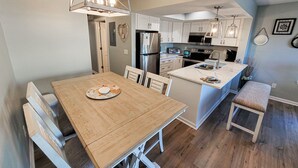A modern kitchen with white cabinets, stainless steel appliances, and a wooden dining table with a bench. - Surf Condos 318 - Ocean Views & Poolside Fun! (Surf City)