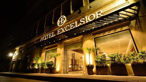 Entrada de un hotel con un cartel que dice "HOTEL EXCELSIOR" y una fachada moderna con grandes ventanales y plantas en macetas.
