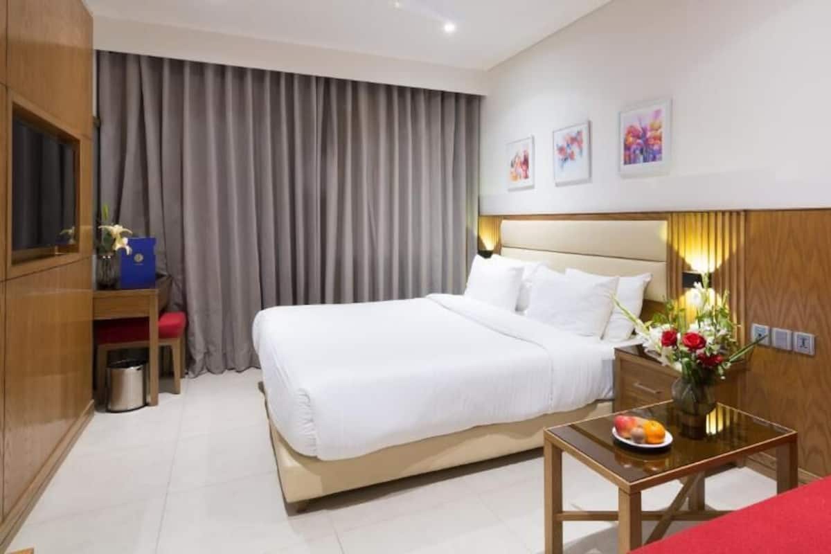 Une chambre d’hôtel avec un grand lit, un bureau agrémenté d’un vase de fleurs, un fauteuil rouge et un téléviseur posé sur une commode en bois.