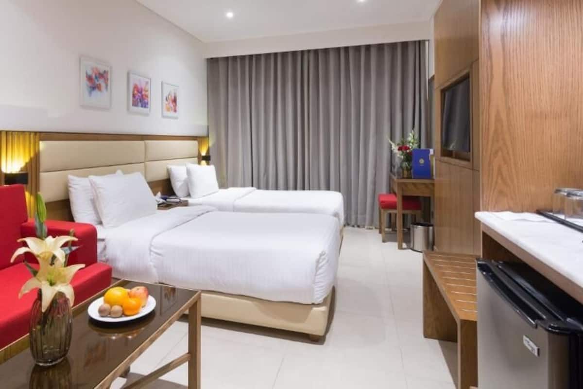 Une chambre d’hôtel avec un canapé rouge, un lit avec du linge de lit blanc, une petite table avec des fruits et un téléviseur posé sur une commode en bois.