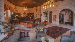 A spacious room with a high ceiling, stone walls, and a mix of seating including sofas and armchairs. A chandelier hangs from the ceiling, and a large rug covers the floor. - Kalsedon Cave Suites (Nevsehir)
