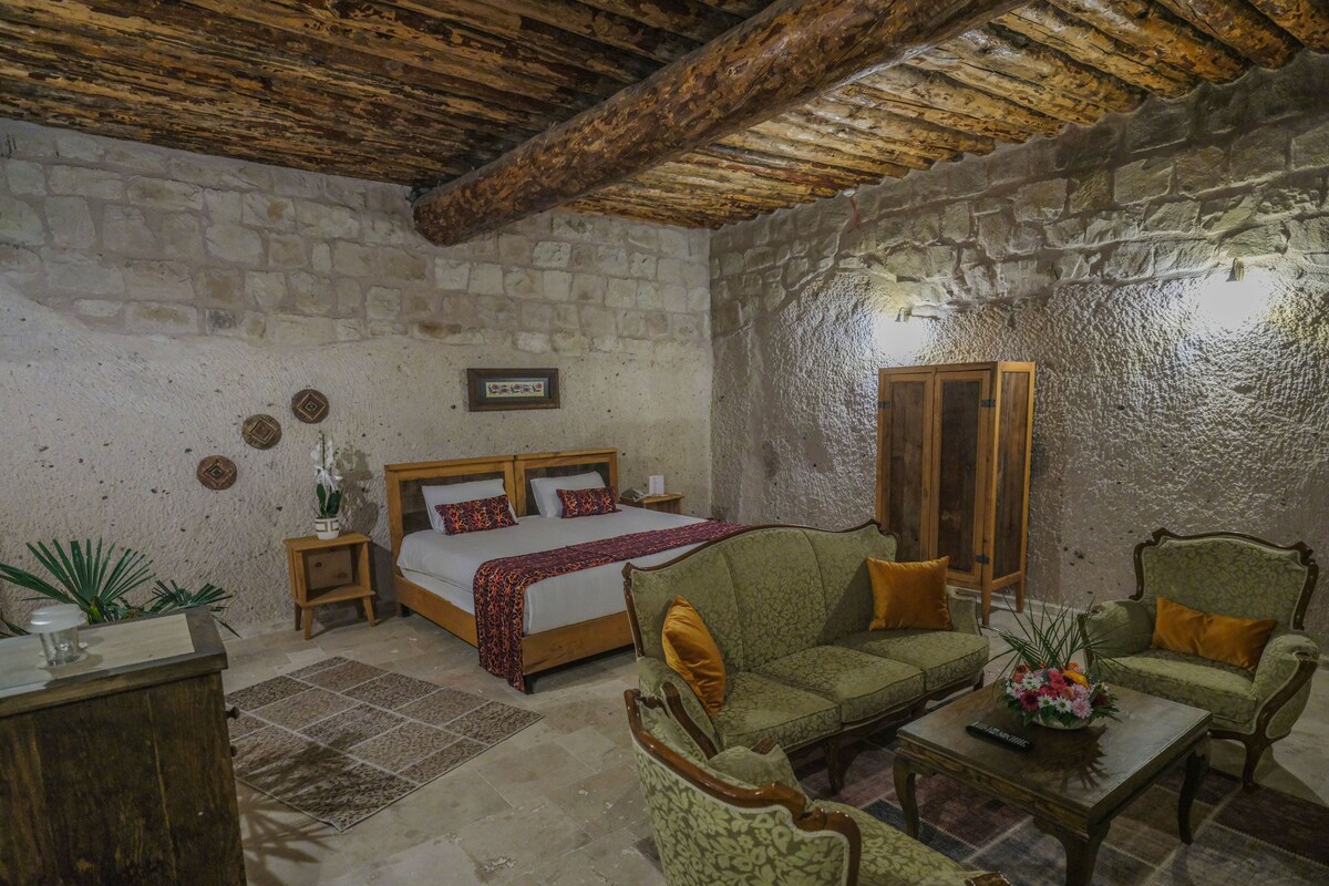 A rustic room with a stone wall, wooden ceiling beams, a bed, a sofa, a chair, a small table, and a cabinet.
