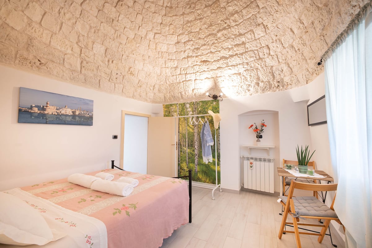 A bedroom with a stone ceiling, a bed with a pink bedspread, a wooden chair, a table with a plant, and a radiator.